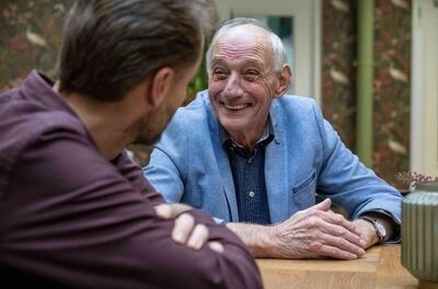 Een oudere man glimlacht en praat met een jongere man aan een houten tafel in een gezellige omgeving.